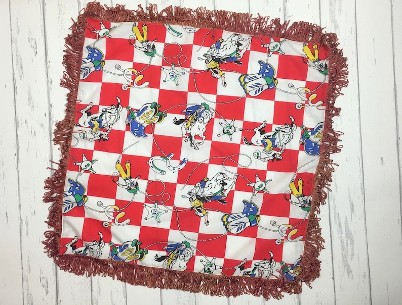 May include: A red and white checkered throw pillow with a fringe border. The pillow features a repeating pattern of cowboys riding horses, stars, and horseshoes.