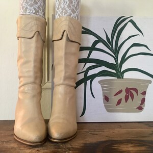 May include: A pair of light beige knee-high boots with a pointed toe and a slight heel. The boots are made of leather and have a fold-over top.