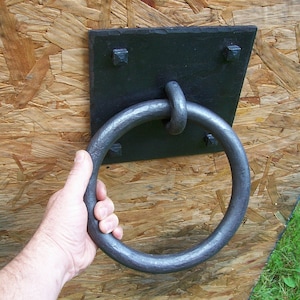 May include: A black metal ring door handle with a square backplate attached to a wooden surface.