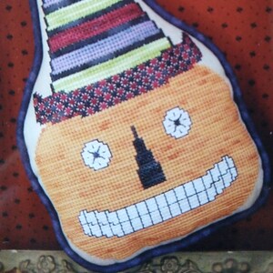 May include: A hand-stitched pumpkin-shaped pillow with a striped hat and a friendly face. The pillow is orange with a black nose and white eyes. The hat is multi-colored with green, purple, red, and white stripes.