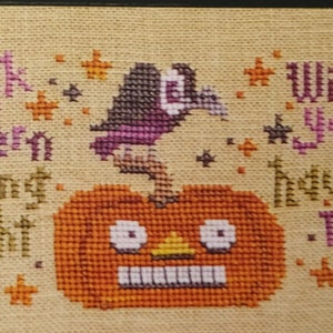 May include: A cross-stitch design featuring a smiling jack-o'-lantern with a purple crow perched on top. The text "Jack lantern smiling bright" is on the left and "Wishing you a haunting night" is on the right.