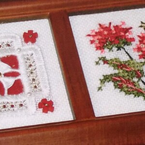 May include: Two framed cross-stitch artworks. One features a red and white geometric design with floral accents. The other depicts red flowers with green leaves and gold star accents. Both are set against a white background.