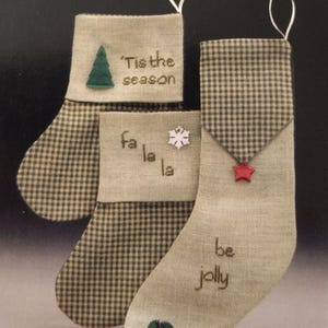 May include: Three Christmas stockings with a green and tan checkered pattern. Each stocking has a different embroidered message: "'Tis the season", "fa la la", and "be jolly". Decorative elements include a green tree, a snowflake, and a red star.