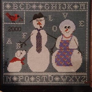 May include: Cross-stitch pattern featuring a snowman couple with a dog. The snowman man is wearing a tie and glasses, the snowman woman is wearing a floral apron, and the dog is wearing a red scarf. The pattern includes the year 2000 and the letters of the alphabet.