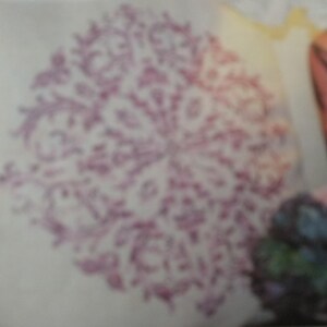 May include: A close-up of a white fabric with a purple floral design. The design is a circular pattern with intricate details.