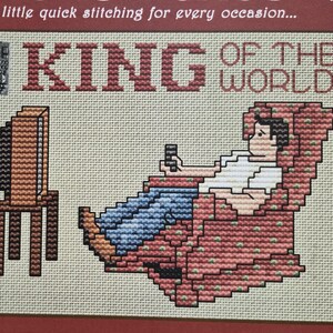 May include: A cross-stitch pattern featuring a person sitting in a red patterned armchair, holding a remote control and facing a brown television set on a stand. The text "KING OF THE WORLD" is stitched in red above the figure on a beige fabric background.