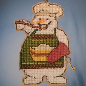 May include: A white snowman wearing a green and red apron and a white chef's hat. The snowman is holding a wooden spoon and a red pot holder. The snowman is holding a key in its right hand.