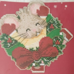 May include: A cross-stitch design featuring a cute mouse wearing a red Santa hat and holding a green wreath with a red bow. The mouse is surrounded by green holly and red berries.