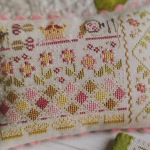 May include: Rectangular decorative pillow with a cross-stitch design. The pillow features a cream-colored background with a floral and geometric pattern in shades of pink, yellow, and green. The edges are trimmed with a pink, scalloped border.