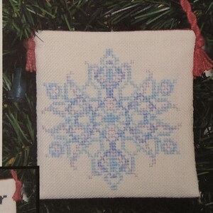 May include: A white fabric ornament with a blue and purple snowflake design. The ornament has a red tassel.