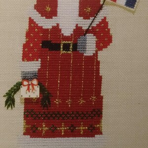 May include: Cross-stitch depiction of Santa Claus holding a French flag. Santa wears a red suit with gold accents, a white beard, and a matching hat. A gift box and the words "Pere Noel" are also visible.