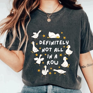May include: Dark gray t-shirt featuring a whimsical design of white cartoon geese and yellow stars arranged around the text "DEFINITELY NOT ALL IN A ROW". The shirt has a vintage, worn look.