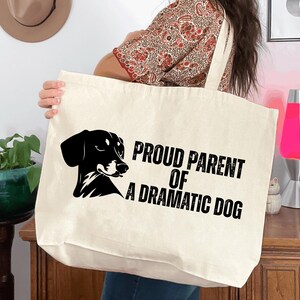 May include: Cream-colored tote bag with a black graphic of a dog and the text "PROUD PARENT OF A DRAMATIC DOG". The bag has a simple design and is held by a person.