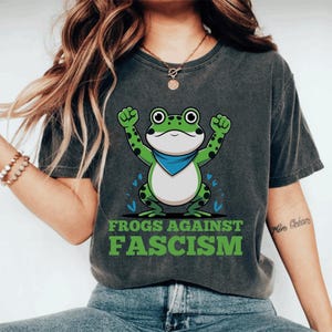 May include: Dark gray t-shirt featuring a cartoon frog with a blue bandana and raised fists. The frog is green with black spots. The text "FROGS AGAINST FASCISM" is printed in green below the frog.