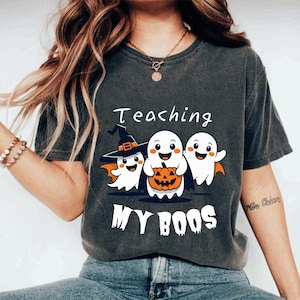 以下が含まれることがあります： 「Teaching My Boos」の文字とハロウィーンをテーマにしたグラフィックがプリントされた濃いグレーのTシャツ。グラフィックには、魔女の帽子をかぶり、カボチャを持っている3人の漫画の幽霊が描かれています。このシャツはカジュアルなスタイルです。