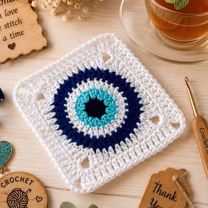 Puede incluir: Un posavasos cuadrado de ganchillo con un motivo central de ojo turco en hilo azul oscuro, azul claro y blanco. El diseño consta de un centro azul oscuro, un anillo azul claro, un borde azul oscuro y un cuadrado exterior blanco con esquinas.