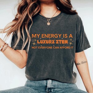 Puede incluir: Camiseta gris oscuro con el texto naranja: "MY ENERGY IS A LUXURY ITEM, NOT EVERYONE CAN AFFORD IT". La camiseta tiene cuello redondo y mangas cortas. El texto está en negrita y estilizado.