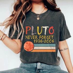 May include: Dark gray t-shirt with the word "PLUTO" in colorful letters. Below, the text reads "NEVER FORGET 1936-2006." A graphic of Pluto and horizontal stripes in orange, white, and blue complete the design. The shirt has a vintage, worn look.