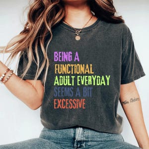 Könnte beinhalten: Dunkelgraues T-Shirt mit dem Text "BEING A FUNCTIONAL ADULT EVERYDAY SEEMS A BIT EXCESSIVE" in bunten Blockbuchstaben. Das Shirt ist ein lässiger Stil, geeignet für den Alltag.