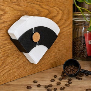 Könnte beinhalten: Ein weißer Papier-Kaffeefilterhalter mit schwarzem Akzent und Korkdetail. Der Halter ist auf einer Holzoberfläche montiert. Kaffeebohnen und ein schwarzer Löffel befinden sich im Vordergrund.