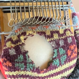 May include: A close-up shot of a sock being knitted on a machine. The sock has a geometric pattern in burgundy, yellow, orange, and green. The knitting machine's needles are visible above the sock. The text "Crafty Creative Co" is visible.
