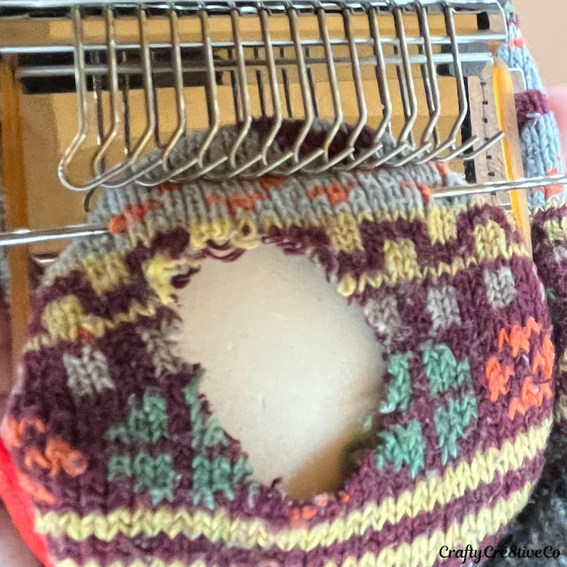 May include: A close-up shot of a sock being knitted on a machine. The sock has a geometric pattern in burgundy, yellow, orange, and green. The knitting machine's needles are visible above the sock. The text "Crafty Creative Co" is visible.