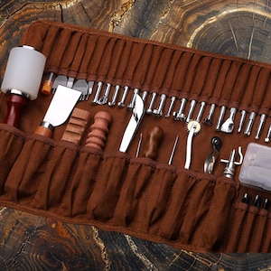 May include: A brown leather roll-up case with a variety of leather working tools, including a hammer, awl, stitching tools, and a small knife. The tools are neatly organized in individual slots.