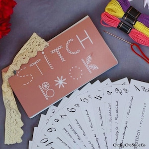 May include: A pink booklet with the word "STITCH" written in white on the cover. The booklet is surrounded by a set of white cards with numbers on them. The cards are labeled "Mini Stitch Book". There are also three spools of embroidery floss in yellow, purple, and pink.