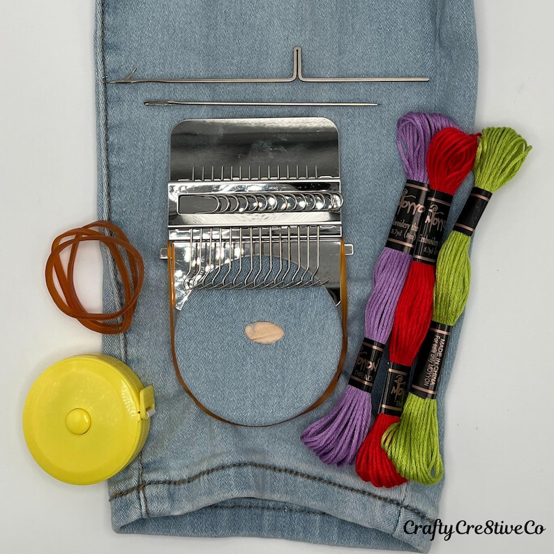 May include: A craft kit featuring a metal weaving tool, needles, and colourful embroidery floss in purple, red, and green. Also includes a yellow tape measure, rubber bands, and a denim-coloured circle.