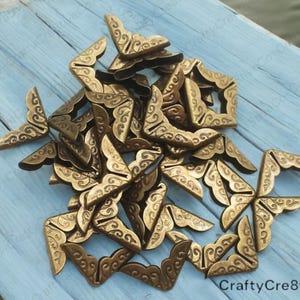 May include: A pile of gold-colored metal corner protectors with a decorative swirl design. The corner protectors are arranged in a random pattern on a light blue wooden surface.