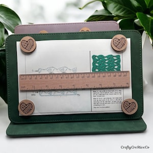 May include: A green folder with a white sheet of paper inside. The paper has a crochet pattern and instructions. There are wooden heart-shaped magnets holding the paper in place. The ruler is 20 cm long.