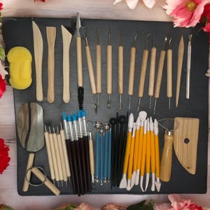 K&ouml;nnte beinhalten: Ein Set mit 30 T&ouml;pfereiger&auml;ten zum Modellieren und Formen von Ton. Die Werkzeuge bestehen aus Holz, Metall und Kunststoff und sind in verschiedenen Formen und Gr&ouml;&szlig;en erh&auml;ltlich. Die Werkzeuge sind auf einer schwarzen Oberfl&auml;che mit einem Schwamm, einem Holzpaddel und einem kleinen Metallschaber angeordnet.