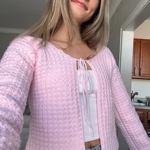 May include: A light pink, open-front crochet cardigan with long sleeves. The cardigan has a textured stitch pattern and a tie closure at the neckline. The garment is worn over a white top.