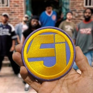 May include: A circular embroidered patch featuring a yellow stylized letter design on a light blue background, enclosed by a thick purple border. The patch is held up against a blurred background of several people standing in front of a brick wall.