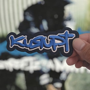 May include: An embroidered patch featuring the word KUSUPT in a stylized blue font with a white outline, set against a dark navy blue background. The lettering has a bold, graffiti-inspired aesthetic with thick stitching and a distinct border.