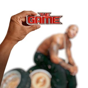May include: An embroidered patch featuring the text THE GAME in bold red letters with a black outline and a white border. The design has a stylized, angular font with a sharp, pointed edge on the right side, creating a distinct graphic emblem.
