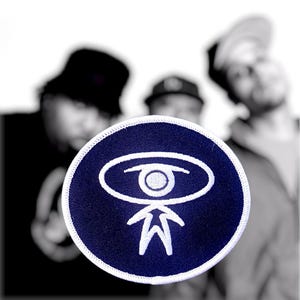 May include: A circular embroidered patch featuring a dark blue background with a white stitched graphic of a stylized figure with an eye-shaped head. The patch has a clean white border edge and is presented against a blurred, out-of-focus background.