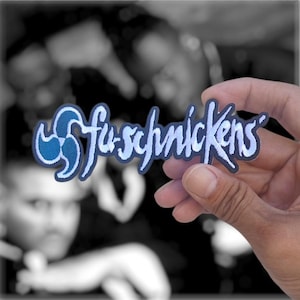 May include: An embroidered patch featuring the text "fu-schnickens" in a stylized, light blue font with a dark blue outline. To the left of the text is a circular, three-pronged emblem in shades of blue. The patch has a dark blue border.