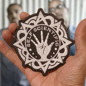 May include: A circular embroidered patch featuring a black background with a white stitched border and a central white hand graphic. The text SELF SCIENTIFIC is arched over the hand, surrounded by a decorative star-shaped geometric frame.