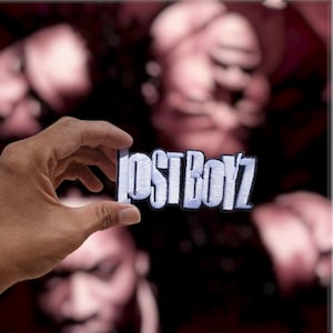 May include: A rectangular embroidered patch featuring the text LOST BOYZ in bold, white, block-style lettering with a thick black border. The embroidery has a visible stitched texture throughout the white letters and the surrounding black outline.