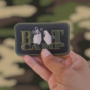 May include: A rectangular patch with the text "BOOT CAMP" in green and white embroidery. The patch features two white dog tags with silver-colored chains. The patch is held in a hand against a camouflage background.