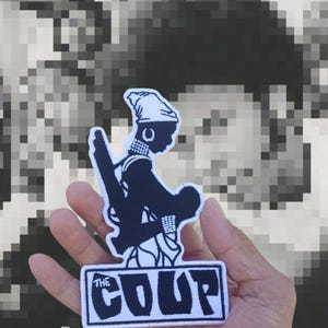 May include: A black and white patch featuring a stylized silhouette of a person with a hat and a weapon. The patch has the text "THE COUP" in a white rectangular border. The background is a pixelated black and white image.