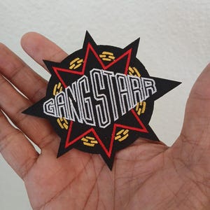 May include: A black embroidered patch featuring a red star shape layered over a circular design with yellow accents. The text GANG STARR is stitched in white across the center of the star. The patch has a sharp, multi-pointed star silhouette.