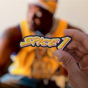 May include: An embroidered patch featuring the text SPICE 1 in a stylized yellow font with a red outline and a thick black border. The lettering has a textured, stitched appearance, creating a bold graphic design suitable for application onto fabric items.