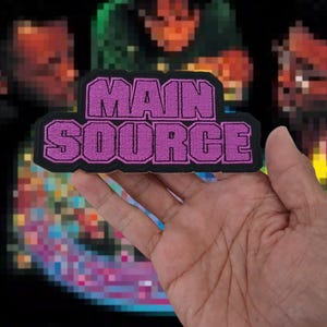 May include: A purple and black embroidered patch with the words "MAIN SOURCE" in a bold, stylized font. The patch is held in a hand, with a pixelated background featuring blurred figures and colors.
