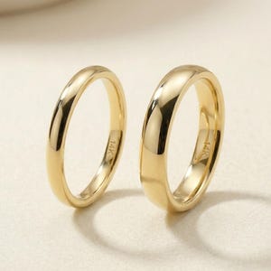 May include: Two polished yellow gold bands with a smooth, rounded profile. Each ring features a clear 14K hallmark stamp on the inner surface. The bands have a high-shine finish that reflects light, showcasing a simple, solid metal construction.