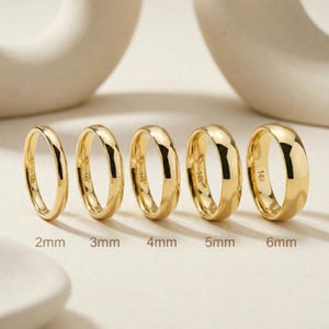 Peut inclure: Cinq alliances en or de largeurs variables, de 2 mm à 6 mm, sont présentées. Chaque bague est polie et reflète la lumière, mettant en valeur un design lisse et arrondi. Les bagues sont étiquetées avec leurs tailles respectives.