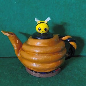 Könnte beinhalten: Eine handgefertigte Keramik-Teekanne in Form eines Bienenstocks mit glänzender orangefarbener Oberfläche. Der Deckel zeigt eine kleine, gelb bemalte Bienenfigur mit weißen Flügeln und schwarzen Details. Der Griff ist dunkelbraun und gedreht.