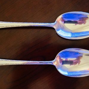 May include: Two silver spoons with decorative handles and a reflective surface. The spoons are placed parallel on a dark brown surface. The spoons are likely for serving or eating, possibly part of a cutlery set.