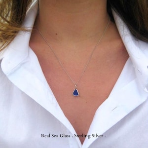 Puede incluir: Un collar que presenta una pequeña pieza de vidrio marino de color azul intenso en forma de lágrima, suspendida de una fina cadena de metal plateado. El colgante está sujeto con una sencilla anilla plateada de diseño refinado.